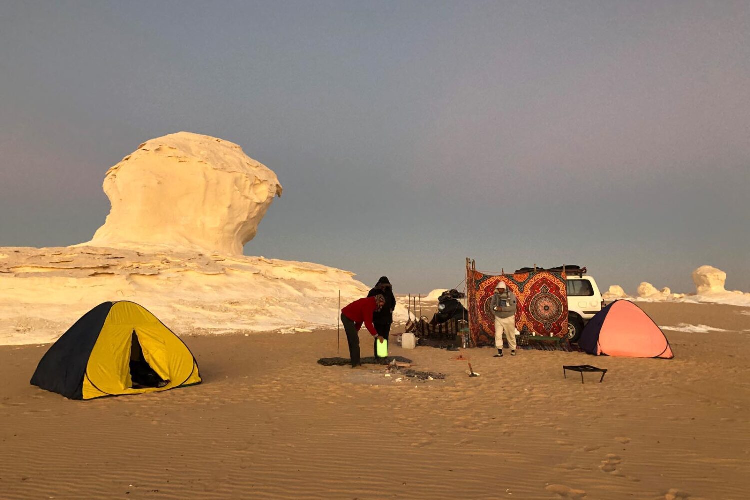 Um acampamento simples no vasto Deserto Branco, com algumas tendas pequenas e veículos 4x4, mostrando a imensidão da paisagem ao amanhecer ou entardecer