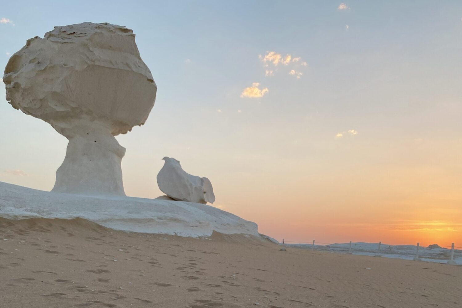 Uma icónica rocha de giz branco em forma de cogumelo, banhada pela luz dourada e suave de um nascer do sol no Deserto Branco do Egito, com sombras alongadas