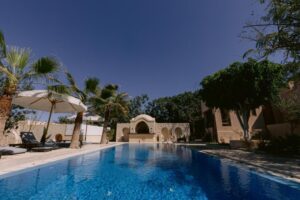A resort swimming pool under a clear blue sky, with palm trees and sun loungers with white umbrellas on the left. In the background