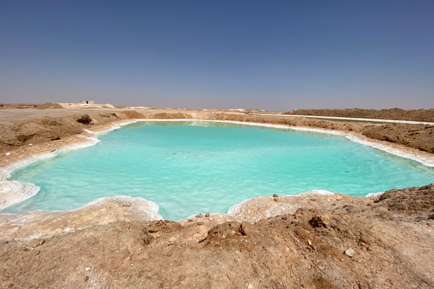 Extensa superfície de um lago salgado de cor turquesa no Oásis de Siwa, com margens brancas de depósitos de sal e a paisagem desértica ao fundo