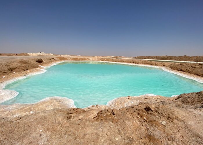 Extensa superfície de um lago salgado de cor turquesa no Oásis de Siwa, com margens brancas de depósitos de sal e a paisagem desértica ao fundo